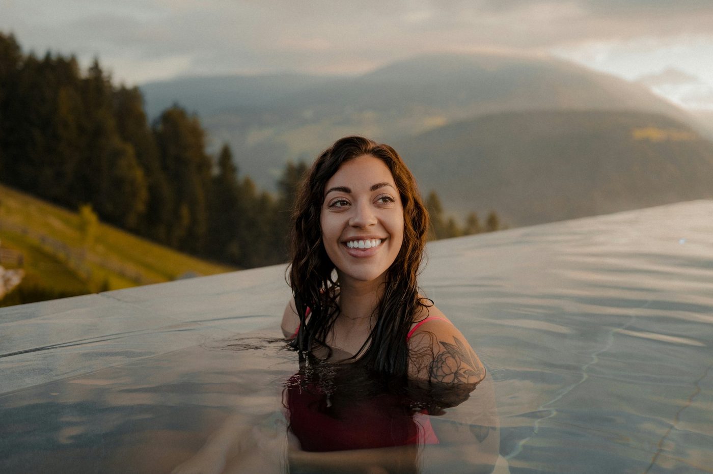A smiling woman in an outdoor pool with forested mountains behind her at sunset.