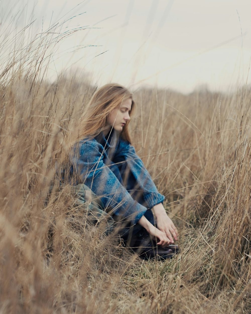 A woman sitting alone in tall dry grass, looking down, lost in thought.